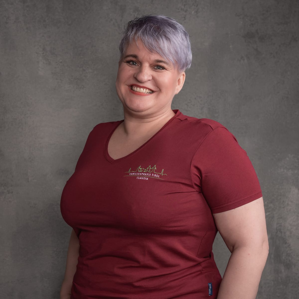 Claudia, Pflegeassistentin in der Familienpraxis Tirol, im bordeauxfarbenen Poloshirt mit Praxislogo, lächelnd vor grauem Hintergrund.