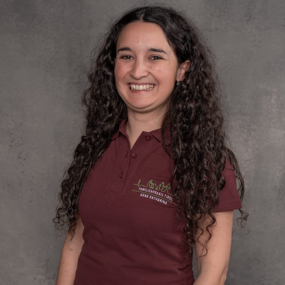 Anna Katharina, Ordinationsmanagerin der Familienpraxis Tirol, im bordeauxfarbenen Poloshirt mit Praxislogo, lächelnd vor grauem Hintergrund.