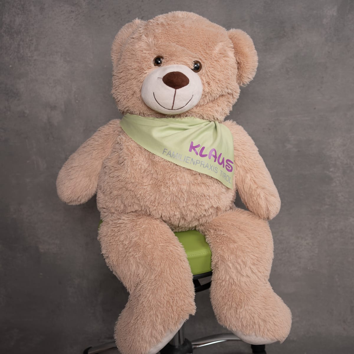 Klaus, das Maskottchen der Familienpraxis Tirol – ein großer beigefarbener Teddybär mit Halstuch und Praxislogo, sitzend auf einem grünen Bürostuhl vor grauem Hintergrund.